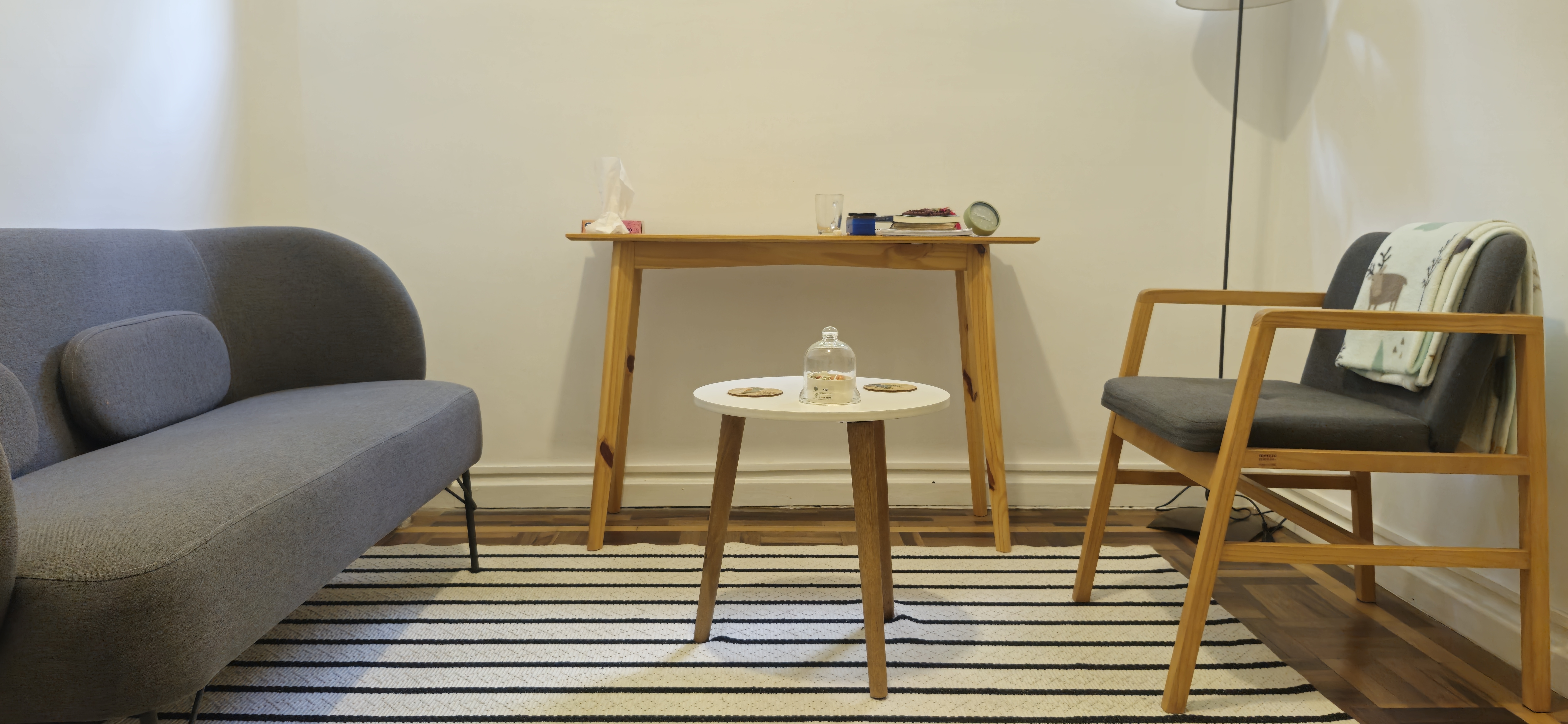 Sala de atedimento individual com sofá cinza, na frente uma cadeira de madeira com estofamento azul, ao chão um tapete listrado azul e branco. Há uma mesinha redonda no centro com porta copos e um aparador de madeira mais ao fundo da foto. Há lustre no canto direito, cortado pela imagem.